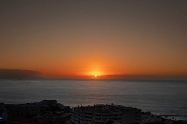 Parlayan turuncu gökyüzü ve sakin su yüzeyi ile okyanusun üzerinde manzaralı bir gün batımı. Önplandaki şehir binalarının silueti kıyı manzarasına kentsel atmosfer ekler. Seyahat, turizm ve romantik kavramlar için ideal..