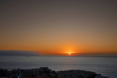 Parlayan turuncu gökyüzü ve sakin su yüzeyi ile okyanusun üzerinde manzaralı bir gün batımı. Önplandaki şehir binalarının silueti kıyı manzarasına kentsel atmosfer ekler. Seyahat, turizm ve romantik kavramlar için ideal..