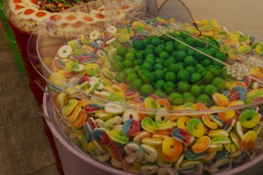 Close up of assorted gummy candies including multicolored rings with sugar coating and bright green sweets displayed in transparent container at candy store. Concept of sweet desserts, childhood, and confectionery.