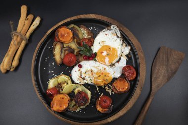 Fried eggs with roasted vegetables including sweet potato, zucchini, tomato and potato slices on black plate. Concept of healthy breakfast, homemade cooking, organic food and gourmet meal