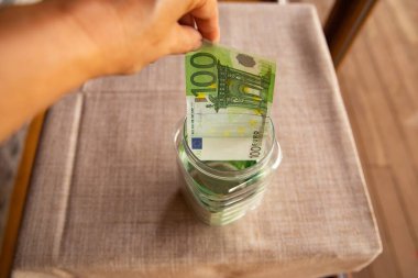 Close up of human hand putting 100 euro banknote into glass jar. Concept of personal savings, financial planning, investment, deposit, and money management.