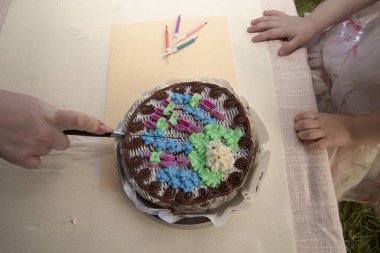 Close up of adult hand cutting chocolate birthday cake with colorful cream decoration while children wait nearby. Concept of family celebration, childhood joy, sweets, and holiday tradition.