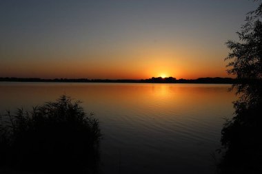 Huzurlu bir yaz akşamı manzarası nehir ve gün batımı yansımaları ağaç siluetleri ile birlikte doğanın rahatlığını ve seyahatini simgeliyor.