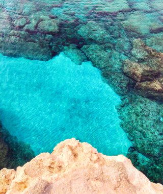 Kayalık, berrak mavi suyla kaplanmış. Lagre taşları suda, dikey üst manzara. Kıbrıs Rum Kesimi 'nin Ayia Napa kenti yakınlarındaki Akdeniz kıyısında. Yaz güneşli manzarası.