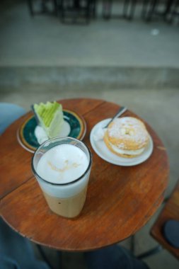 Kremalı buzlu latte, bir dilim yeşil çay krepli kek ve kremalı pasta ile ahşap bir kafe masasının yukarıdan görünüşü. Kahve molası, tatlı zamanı ya da dinlendirici kahve anları için mükemmel bir ortam..