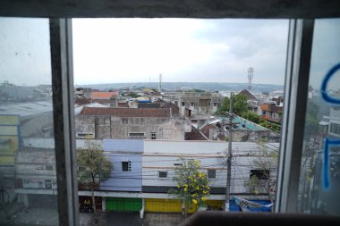 Şehir sokak manzarası, renkli kapalı dükkan kapılarını, çiçekli ağaçları ve kiremit çatılı eski beton binaları gösteriyor. Güç hatları ve iletişim kuleleri cesur şehir atmosferine ekleniyor.