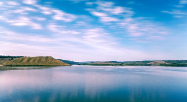 panoramik görkemli manzara kırsal tepeler ve sessiz gölün ön plandaki düz su yüzeyi, gün batımından önce yumuşak renklerde bulutların bulanık hareketi, bahar mevsimi