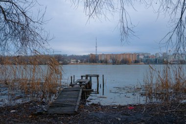 depressive ugly third world country side Eastern Europe outskirts river shore line with ruined wooden pier and background cement buildings in gray low light and colors environment clipart