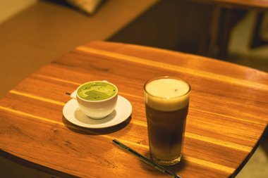 Sıcak bir kafe deneyimin sıcaklığını ve cazibesini yakalayan, ahşap bir masada zarif bir şekilde sergilenen, uzun bir bardak ferahlatıcı buzlu kahveyle eşleştirilmiş bir fincan kremalı matcha latte..