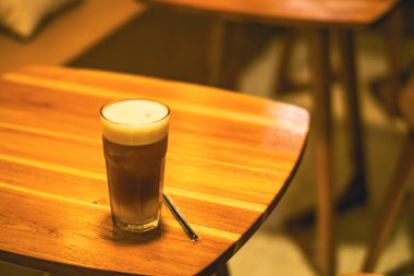 Sıcak bir kafe deneyimin sıcaklığını ve cazibesini yakalayan, ahşap bir masada zarif bir şekilde sergilenen, uzun bir bardak ferahlatıcı buzlu kahveyle eşleştirilmiş bir fincan kremalı matcha latte..