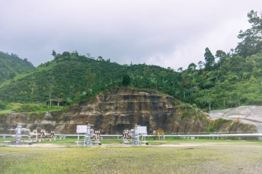 Verimli tepelerin ortasındaki bir jeotermal tesiste gelişmiş boru tesisleri, dikey depolama ve güvenlik ve sürdürülebilirlik için araziyi güçlendiren işçiler ile eğim dengelenmesi gösteriliyor..