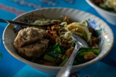 Mie Ayam Bakso 'nun yakın çekimi, tavuk, köfte, yeşil sebze ve kızarmış arpacık soğanlı Endonezya eriştesi. Lezzetli tatların ve dokuların hoş bir karışımı canlı mavi bir masada..