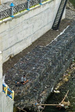 Kirlenmiş bir nehir kıyısında beton duvarlar ve enkazlarla çevrili bir Gabion yapısında tek başına oturan bir adam. Sahne, kentsel zorluklar arasında sessiz bir anı yakalıyor..