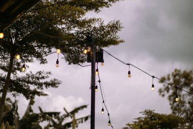 Kırsal bir kutuptan sarkan parlak ip ışıkları bulutlu bir gökyüzünün altında geceyi aydınlatıyor. Sıcak ampuller karamsar atmosferle tezat oluşturarak rahat ve davetkar bir açık hava ortamı yaratıyor..