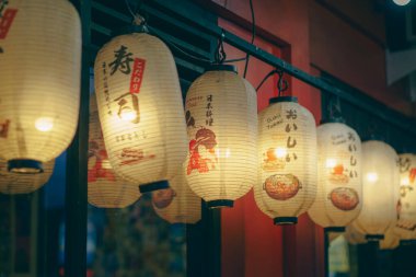 Geleneksel Japon kağıt fenerlerinden oluşan sıra, akşam caddesini aydınlatan sıcak bir şekilde parlıyor. Karmaşık tasarımları ve yumuşak ışıkları Japon gece hayatını çağrıştıran davetkar bir ortam yaratıyor..