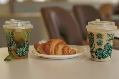 Sıcak bir buzlu kahve, yeşil çay ve beyaz bir masada taze kruvasan. Rahatlatıcı bir kafe anı için mükemmel..
