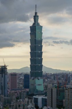 Uzun, modern bir gökdelen alacakaranlıkta bulutlu bir gökyüzünü deler, katmanlı tasarımı hafifçe parlar. Yapı çimenli bir tepenin üzerinde yükselir, uzak dağlar belli belirsiz görünür..