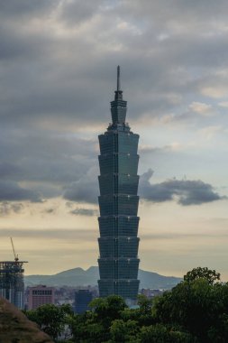 Uzun, modern bir gökdelen alacakaranlıkta bulutlu bir gökyüzünü deler, katmanlı tasarımı hafifçe parlar. Yapı çimenli bir tepenin üzerinde yükselir, uzak dağlar belli belirsiz görünür..
