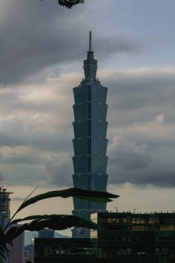 Uzun, modern bir gökdelen alacakaranlıkta bulutlu bir gökyüzünü deler, katmanlı tasarımı hafifçe parlar. Yapı çimenli bir tepenin üzerinde yükselir, uzak dağlar belli belirsiz görünür..