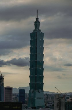 Uzun, modern bir gökdelen alacakaranlıkta bulutlu bir gökyüzünü deler, katmanlı tasarımı hafifçe parlar. Yapı çimenli bir tepenin üzerinde yükselir, uzak dağlar belli belirsiz görünür..