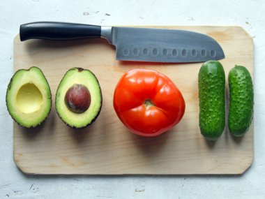 Bir taş, bir domates ve bir salatalık ile bir avokado mutfakta bir bıçağın yanında, tahta bir tahtanın üzerinde beyaz bir masanın üzerinde yatıyordu. Temiz sebzeler, çiftçilik, salata pişirme, köy mutfağı, sağlıklı yiyecekler.