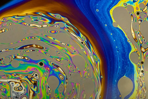 Bright, vibrant purple, blue, orange and sepia pastel colours swirl around intense black dots in a beautiful colorful pattern on the surface of a soap bubble. 