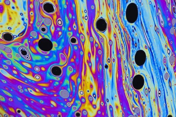 Bright green, pink, purple, blue and orange pastel colours swirl around intense black and white dots patterns on the surface of a soap bubble. 