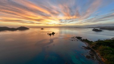 Beautiful clouds obscuring the sun rising over the beach town of Oakura, New Zealand. Silhouetted rocky islands and outlets frame the serene and tranquil scene clipart