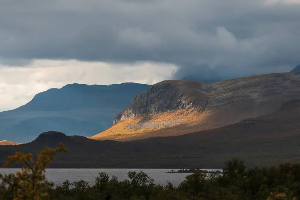 Sunny mountain side. Cloudy weather otherwise creating moody atmosphere. Lakeshore on the foreground. Norway region.