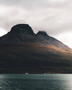 Görkemli dağın eteklerine bakan küçük evler. Karamsar atmosfer. Dağ yamacında güneş ışığı. Dikey fotoğraf.
