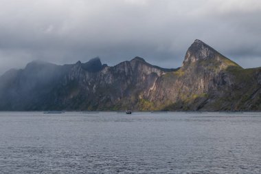 Uçurumun kenarından Segla ve Hesten dağlarının güzel manzarası. Dağları aydınlatan güneş. Denizdeki balıkçı teknesi ve somon çiftlikleri. Norveç 'teki Senja Adası.