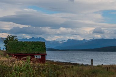 Çimenlerle kaplı, deniz kenarında kırmızı, küçük bir bina. Arka planda inanılmaz bir dağ manzarası var. Gökyüzünde güzel bulutlar.