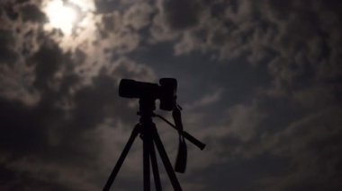 Camera on tripod under night cloudy sky with moon
