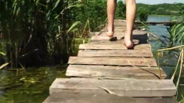 Çıplak ayaklı bir çocuk yazın nehir kıyısında tahta bir köprü boyunca yürür..