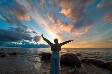 Gün batımında deniz kıyısında ellerini kaldırmış güzel bir kadının silueti.