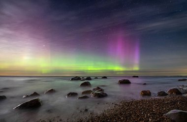 Kuzey ışıklarıyla yıldızlı bir gökyüzünün altında kayaları olan deniz manzaralı bir yer..
