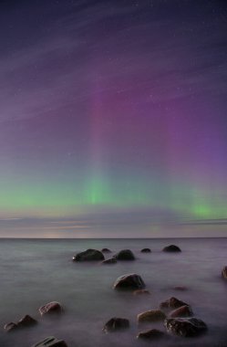 Kuzey ışıklarıyla yıldızlı bir gökyüzünün altında kayaları olan deniz manzaralı bir yer..