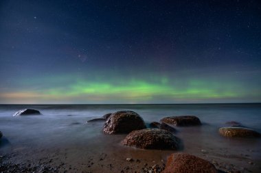 Kuzey ışıklarıyla yıldızlı bir gökyüzünün altında kayaları olan deniz manzaralı bir yer..