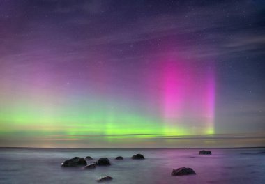 Kuzey ışıklarıyla yıldızlı bir gökyüzünün altında kayaları olan deniz manzaralı bir yer..