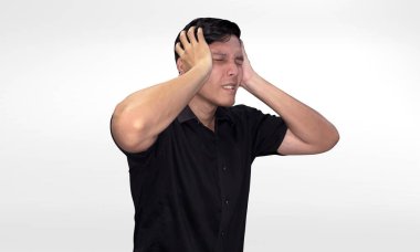 Ayakta duran siyah tişörtlü genç bir adam baş ağrısı çekiyor, acı çektiğini gösteren bir yüz ifadesi var, iki elinde başını tutuyor, beyaz arka planda izole edilmiş..