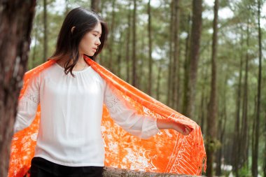 Ormandaki genç Asyalı kadın beyaz bluz giyerken sırtında turuncu Tai Dai elbisesi taşıyor. Doğa fotoşopu.