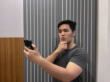 Handsome young man taking selfie using phone camera while posing, hand on chin with rizz facial expression. Narcissistic personality behavior, self obsessed clipart