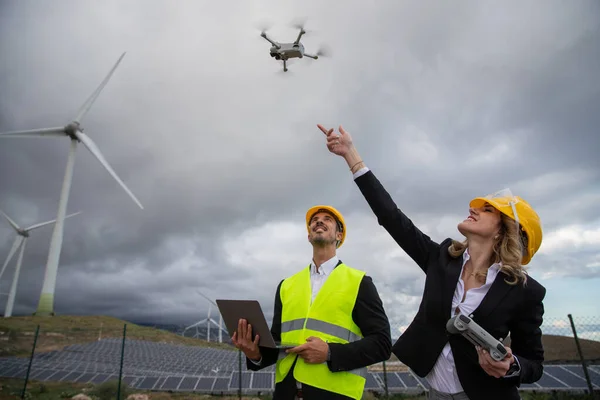 Two engineers at work fly a drone and use the laptop in a solar farm with wind turbines