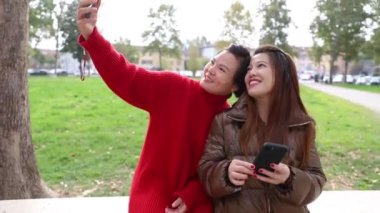 İki Çinli kız arkadaş parkta bir bankta otururken selfie çekiyorlar.