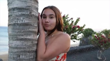 Düşünceli bir genç, deniz kenarındaki bir palmiye ağacına yaslanır ve etrafına bakar.