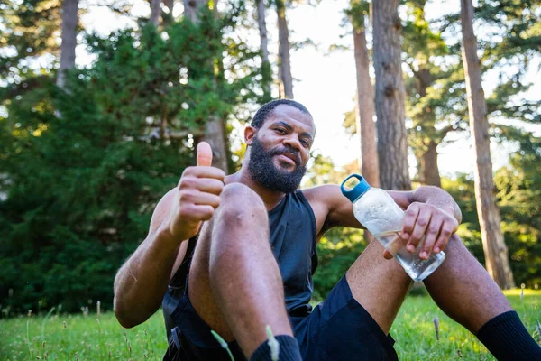 Elinde Bir Şişe Suyla Çimenlikte Oturan Bir Sporcu Başparmağını Kaldırır — Stok fotoğraf