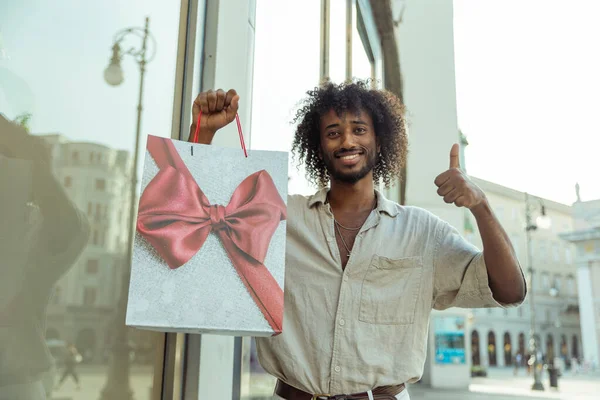 A man goes shopping and is happy, he gives the thumbs up with the shopping bag