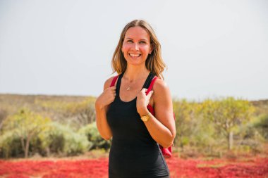 Bir kadın gülümsüyor ve siyah bir atlet ve kırmızı bir sırt çantası giyiyor. Kırmızı çiçeklerle dolu bir tarlada duruyor.