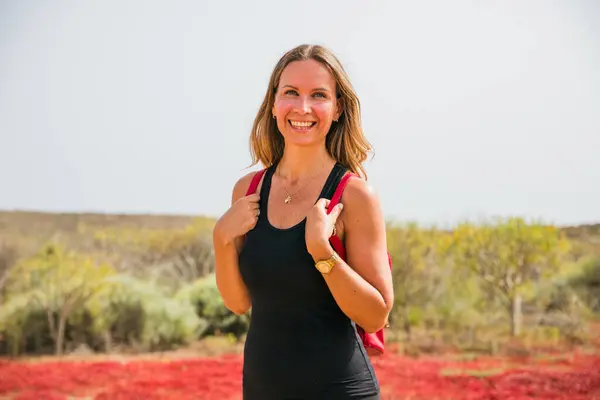 Bir kadın gülümsüyor ve siyah bir atlet ve kırmızı bir sırt çantası giyiyor. Kırmızı çiçeklerle dolu bir tarlada duruyor.