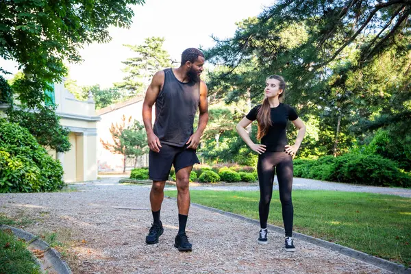 A man and a woman are walking on a path in a park. The man is wearing a black tank top and shorts, while the woman is wearing a black shirt and black pants